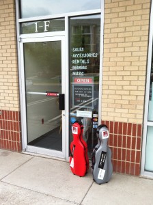 Photo of the red and the gray: twin cello-shaped viola cases set on the sidewalk in front of Lashof Violins in Gaithersburg, Maryland.