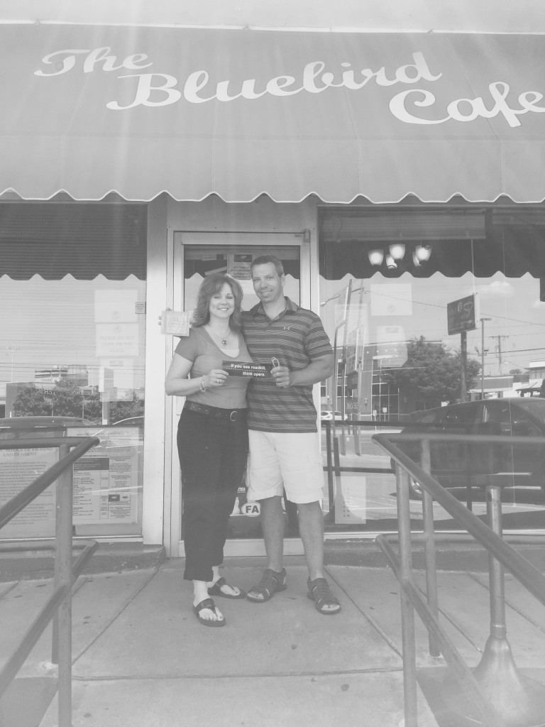 Black and white photo of a couple standing in front of the Bluebird Cafe holding a CD of A Roadkill Opera and a bumper sticker that reads "If you see roadkill, think opera"