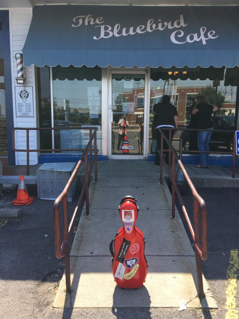 Photo of red viola case in foreground as people peer in the windows of the Bluebird Cafe. Stephan Alexander Parker can be seen in the reflection, taking the picture