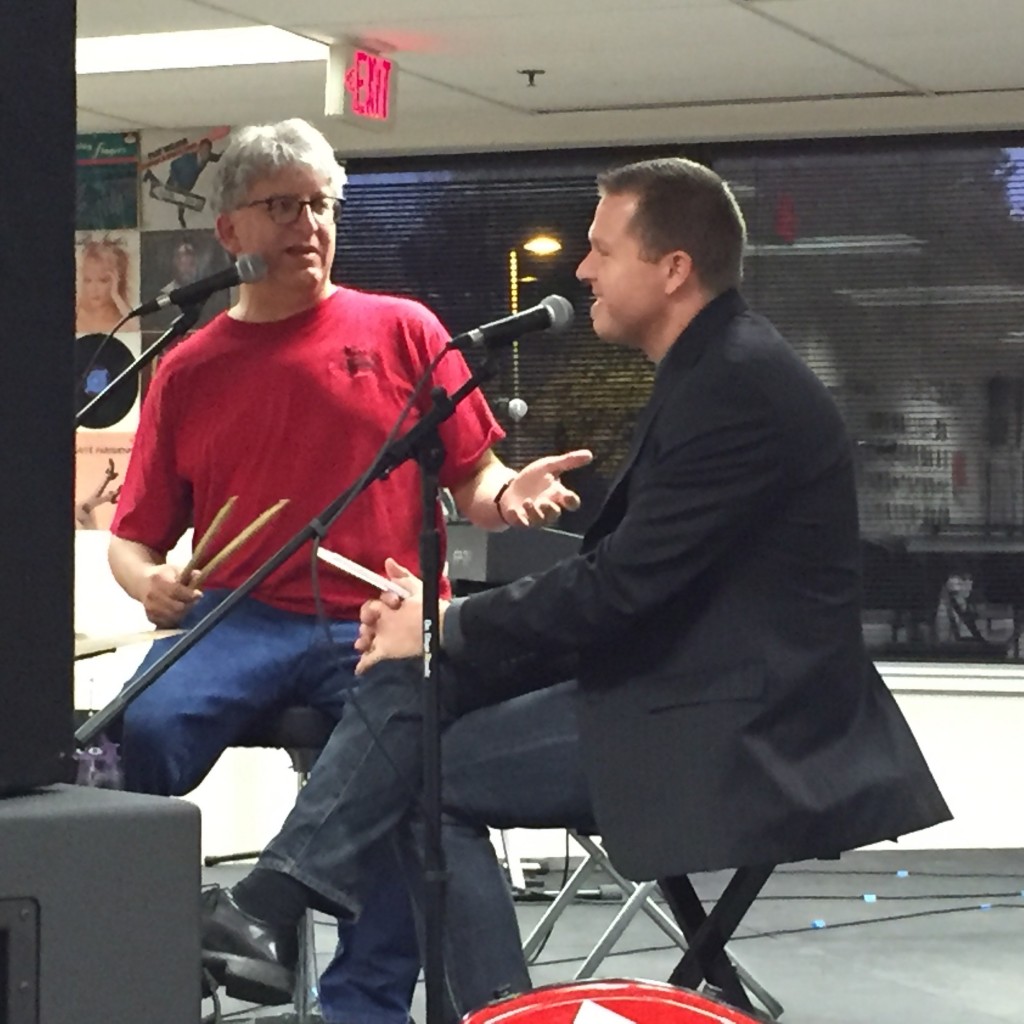 Photo of Parker gesturing to Dokken while sitting at microphones. Parker is holding drumsticks.