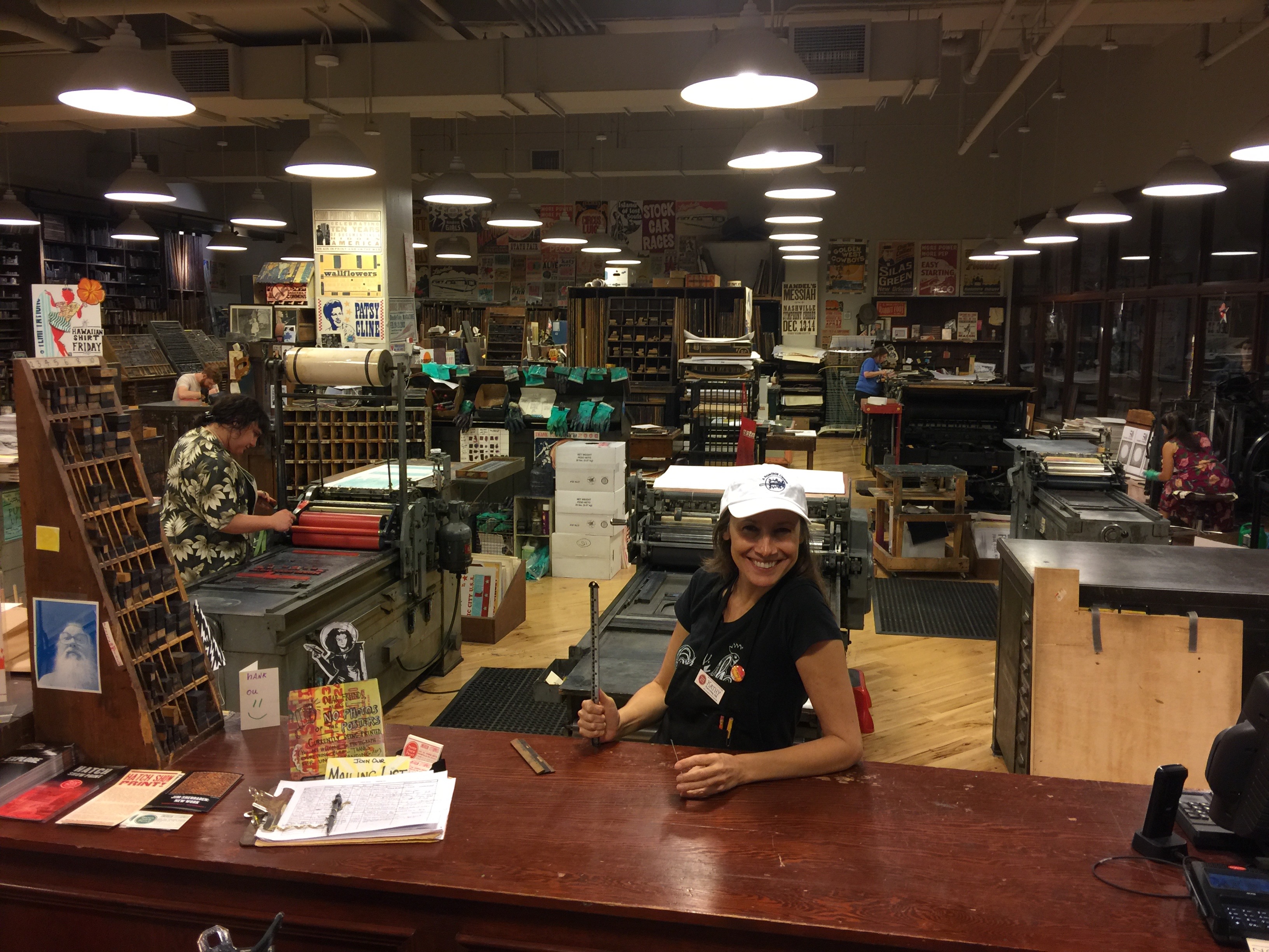 Photo of Kathy standing at service desk in front of the Hatch Show Print workshop full of presses