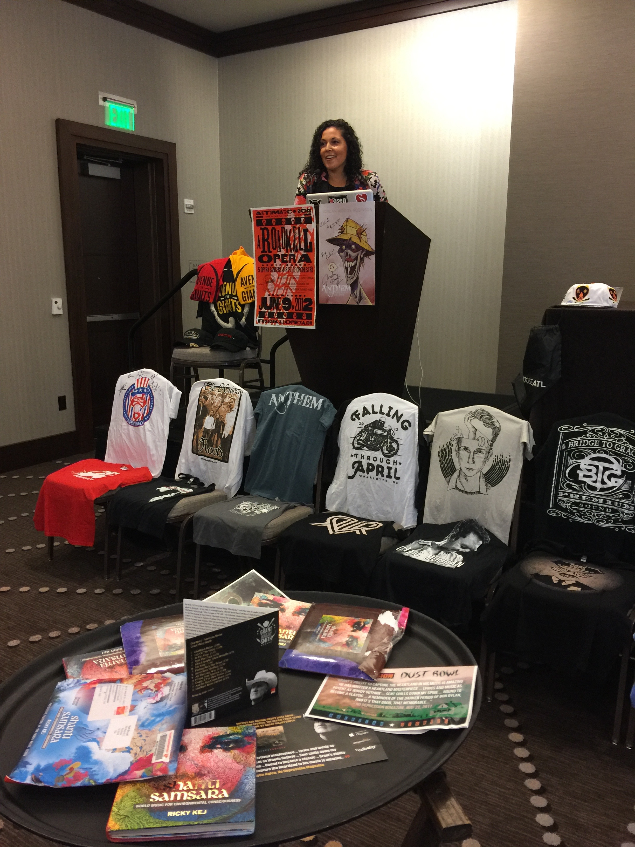 Photo of Vanessa Ferrer at the podium festooned with posters from a Walking Dead star and A Roadkill Opera. In front of the podium is a row of chairs with t-shirts and table vocoder with alternative swag, such as custom incense packaging from a reggae artist.