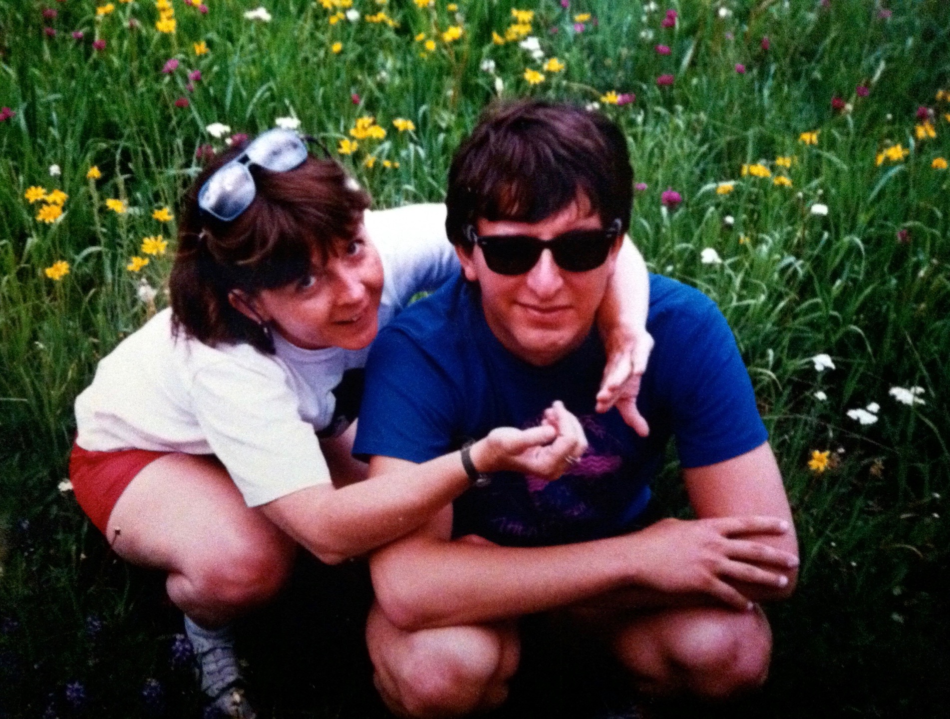 Photo of Debby and Stephan squatting in a field of wildflowers. They are wearing sunglasses and smiling.