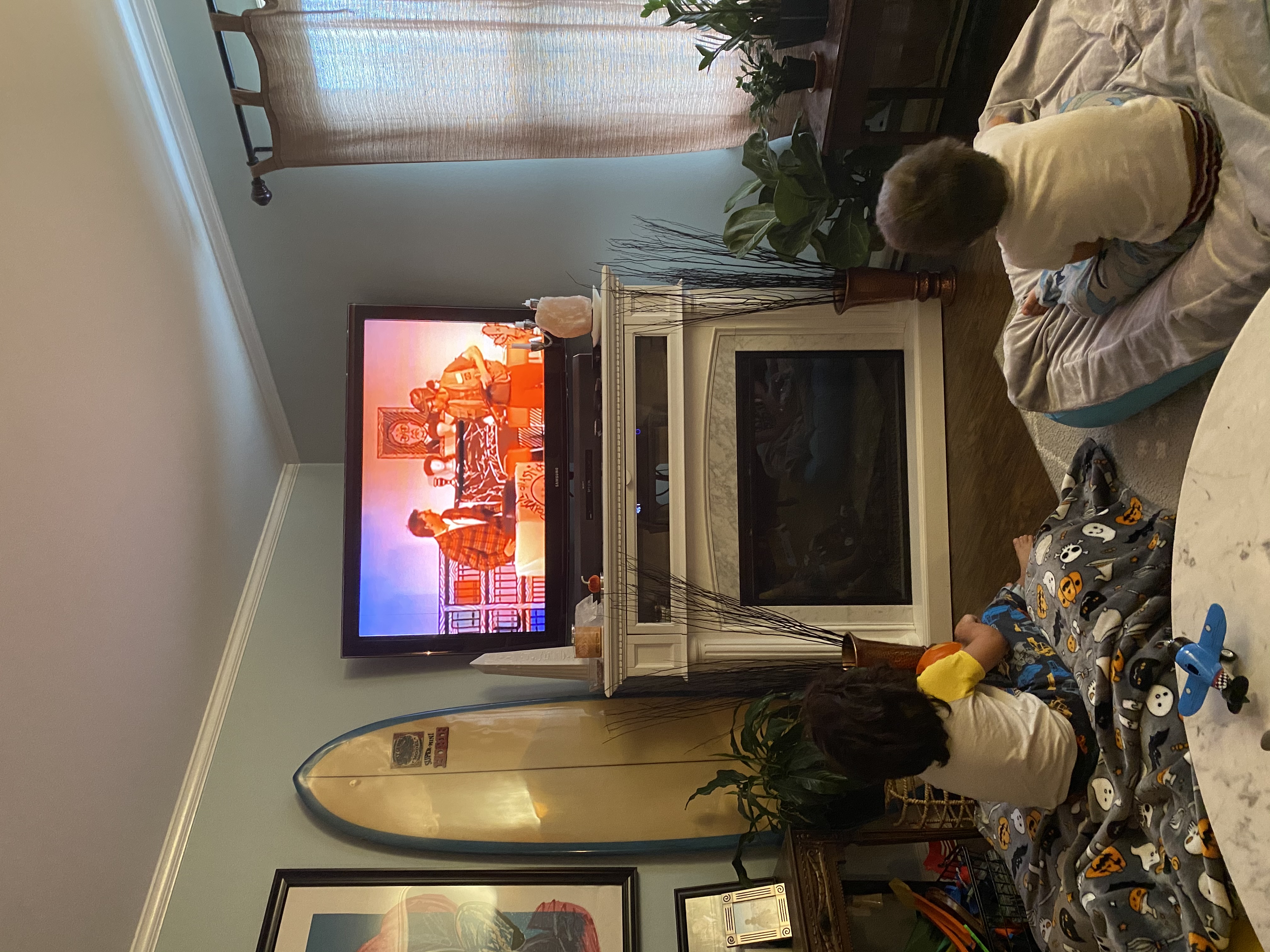 Photo of 3 and 5 year olds watching the livestream on a television, while they sit on the floor of a beach house (based on the surfboard next to the television)