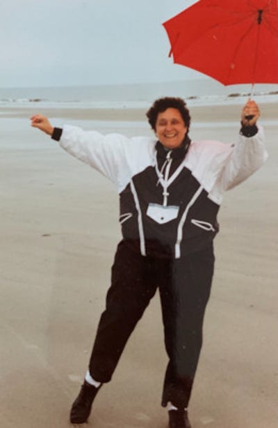 Photo of Elaine Parker, pictured here on the beach in a black and white windbreaker suit and sporting a red umbrella like a tightrope walker dancing atop the wire.